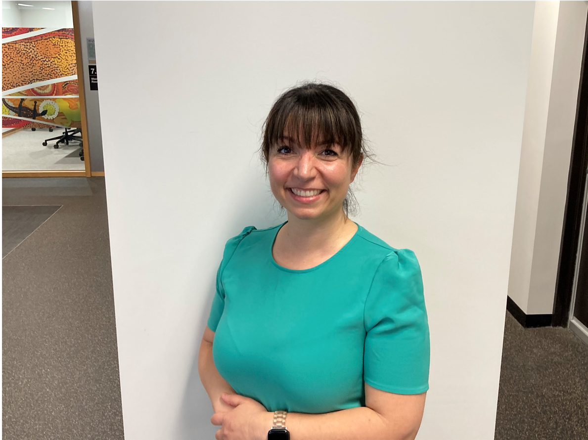 Emmie is wearing a bright green shirt and is standing infront of a white wall. Her brunette hair is tied into a low bun a front fringe. Her arms are crossed across her waist and she is smiling