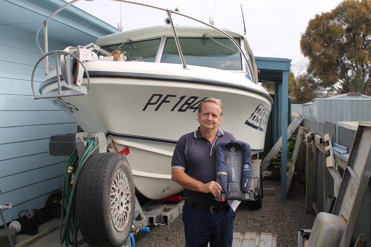 A man standng in front of a boat on a trailer . The man is holding a lifejacket.