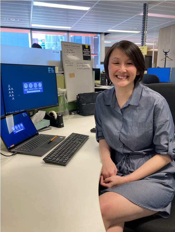 June is sitting at her desk with two computer screens behind her. She is wearing a blue-grey collared dress and her short black hair is straight. She is smiling